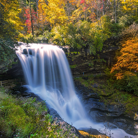 Autumn forest surrounds a wide waterfall cascading over rocky ledges into a dark pool below, with colorful fall foliage framing the scene.