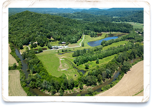 Aerial view of a rural landscape with a winding river, green fields, trees, a small pond, and a few buildings nestled among the greenery.