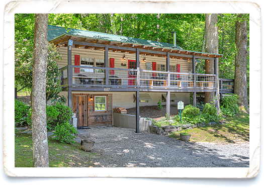 Two‑story rustic cabin in the woods with a covered upper deck and red shutters.