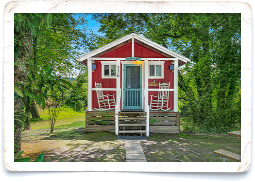 Red wooden cottage with a blue front door and white trim, front porch featuring two rocking chairs among green trees.